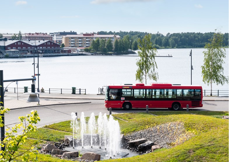 Bild på en röd LLT buss som kör igenom rondellen vid Kulturens hus i Luleå. Mitt i rondellen syns grönska och en fontän i en dam. Bakom bussen syns vattnet i Norra hamn och Norrbottensteatern.