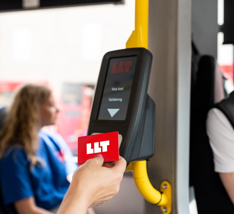 Bild på en kvinnas hand som validerar sitt röda resekort i validatorn ombord på bussen. I bakgrunden syns en ung kvinnlig resenär som tittar ut genom bussfönstret.