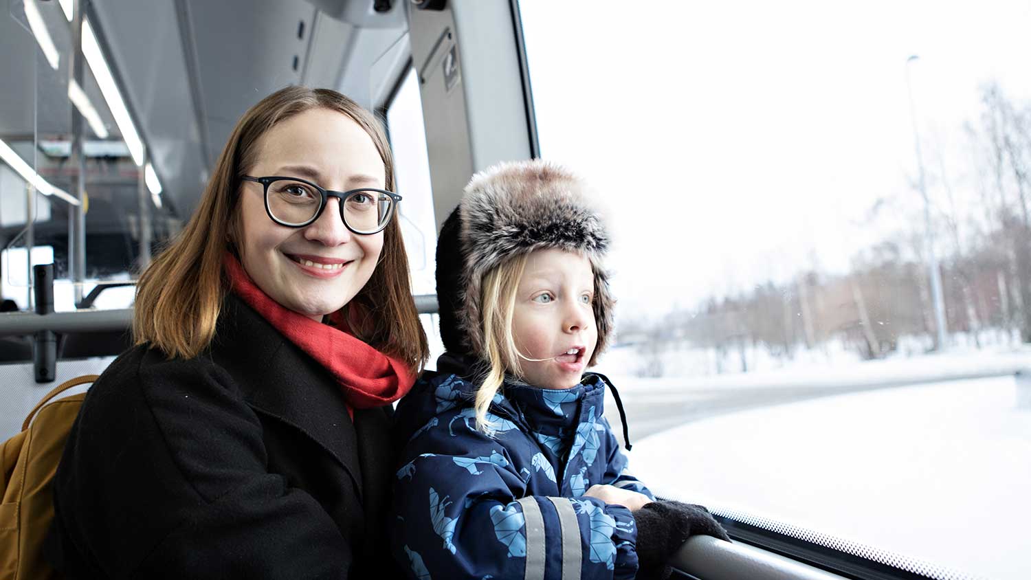 Närbild på mamma och barn som sitter på buss. Mamman tittar in i kameran och barnet tittar ut genom fönstret.