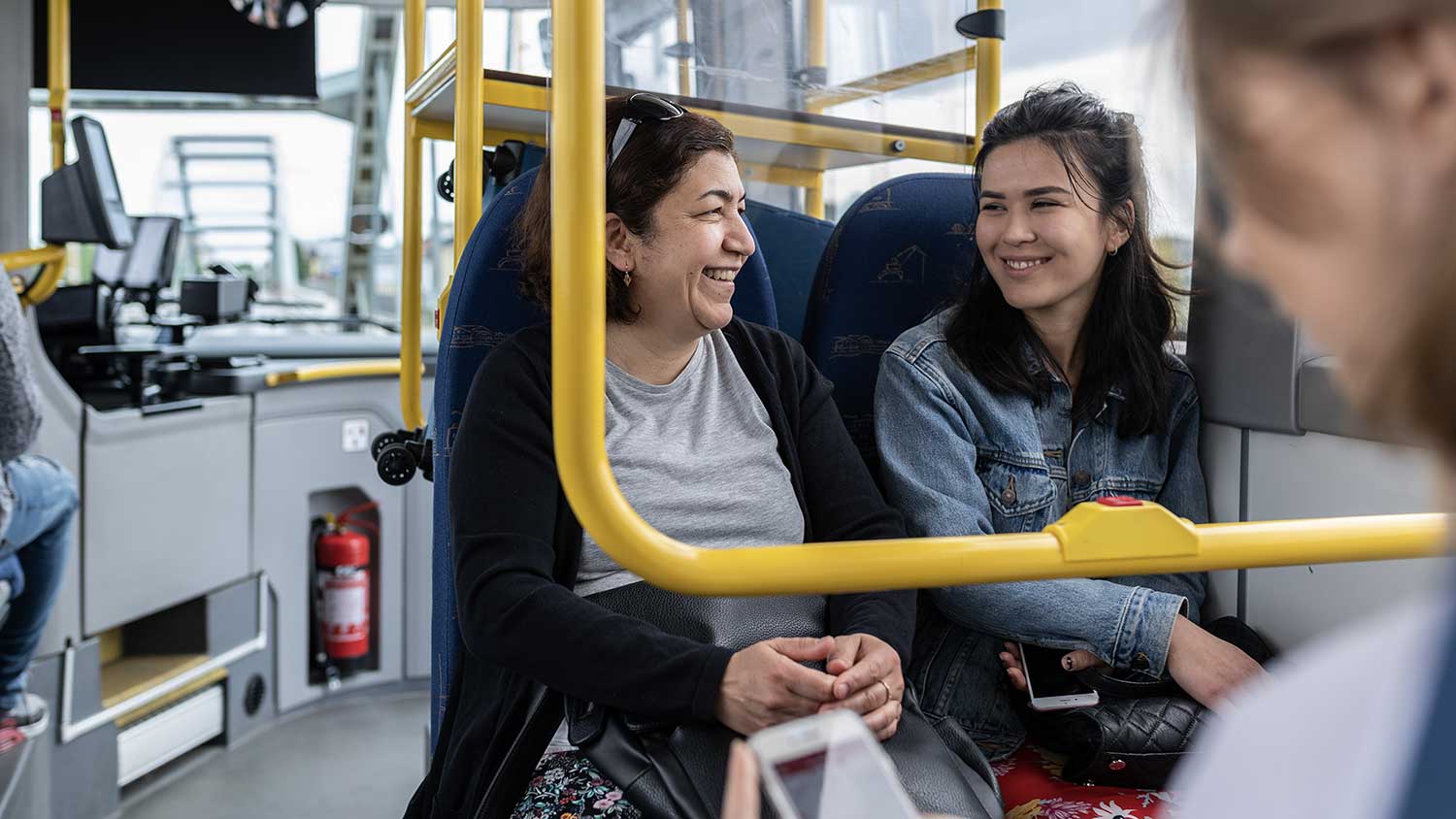 Äldre och yngre kvinna sitter bredvid varandra på buss, tittar på varandra och ler.