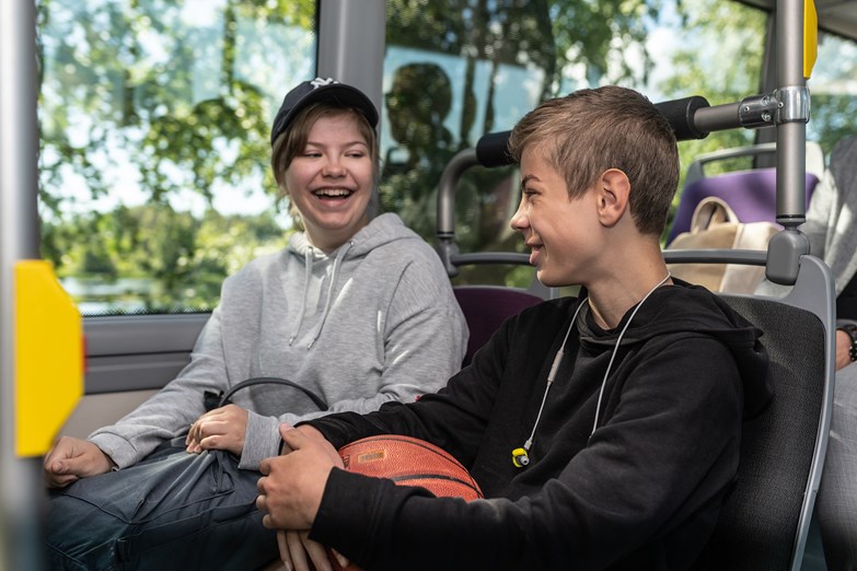 Bild på två glada ungdomar som sitter i en buss. De skrattar och tittar på varandra. Han håller i en basketboll och hon håller i en väska.